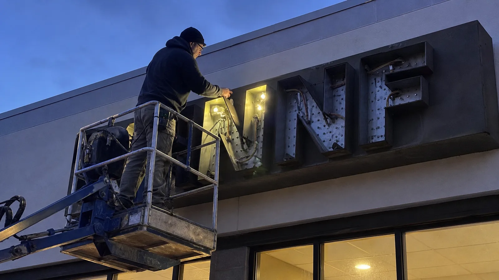 Sign technician inspecting illuminated channel letters from a lift at dusk
